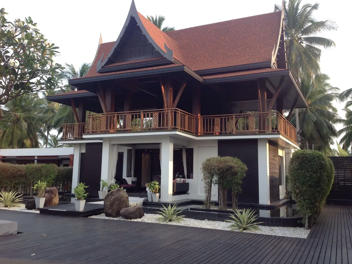 เรือนไม้ทรงไทยยกใต้ถุน หลังคาจั่วสีแดง มีต้นไม้รอบบ้าน